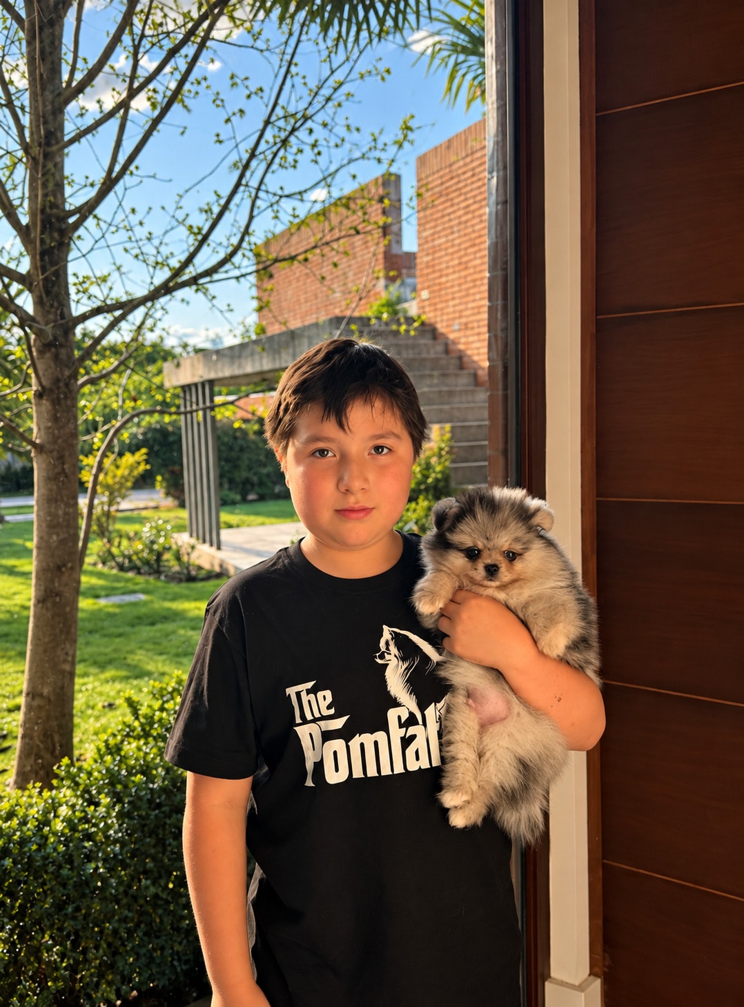 Niño con una polera negra que sostiene en brazos a un cachorro de Pomerania, en la entrada de una casa con jardín